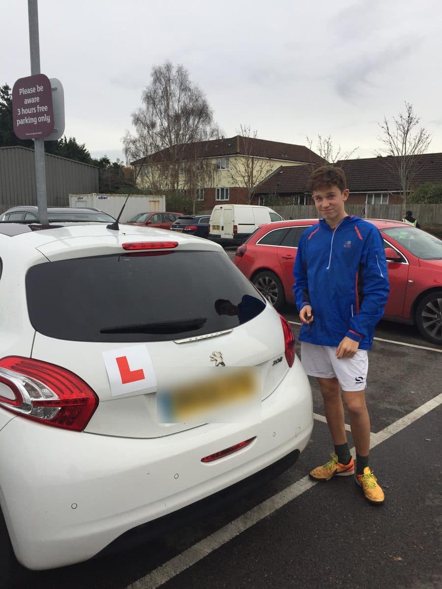 A teenager in a blue jacket standing beside a white learner car in a supermarket car park, car keys in his hand.