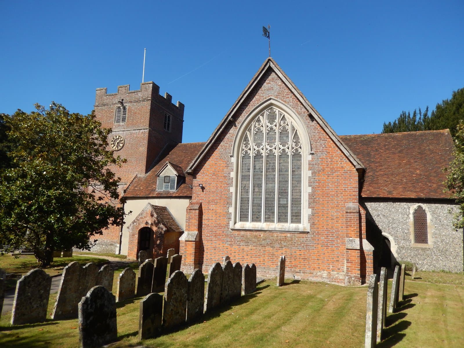 St James' Church, Bramley, from the churchyard, its brick tower with a clock behind the flint and brick east end and a tall traceried window, gravestones in the grass in front.