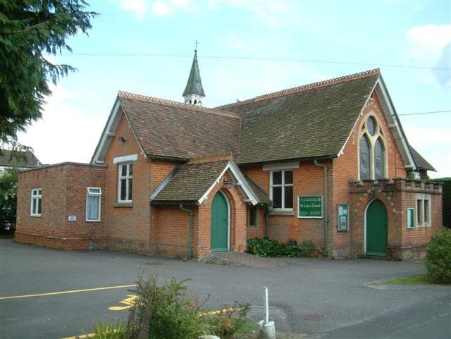 St Luke's Church, Pamber Heath, a red brick Victorian church with a tiled roof, a small spire and two green doors, a conifer to one side.