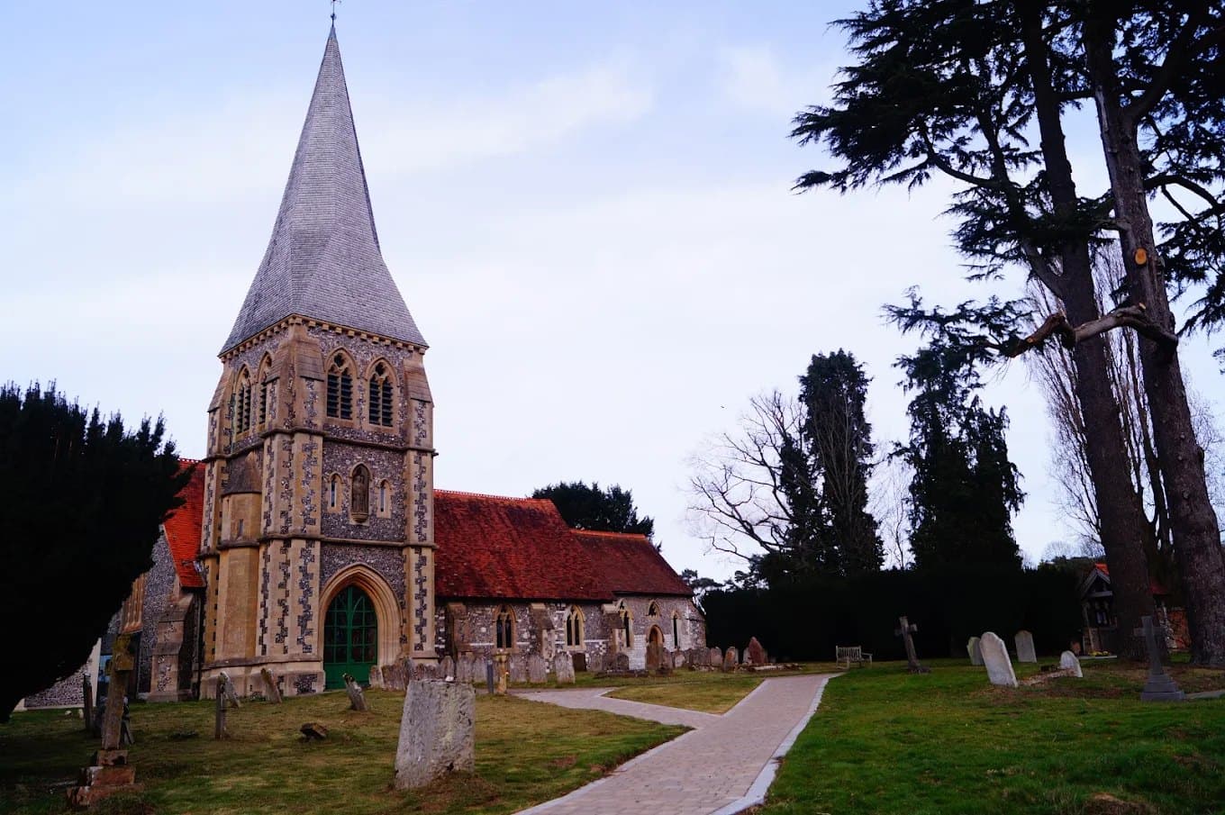 St Leonard's Church, Sherfield on Loddon, a flint church with stone dressings and a tall shingled broach spire over the tower, a red tiled roof, green doors and a paved path through the churchyard, with a cedar and yews behind.