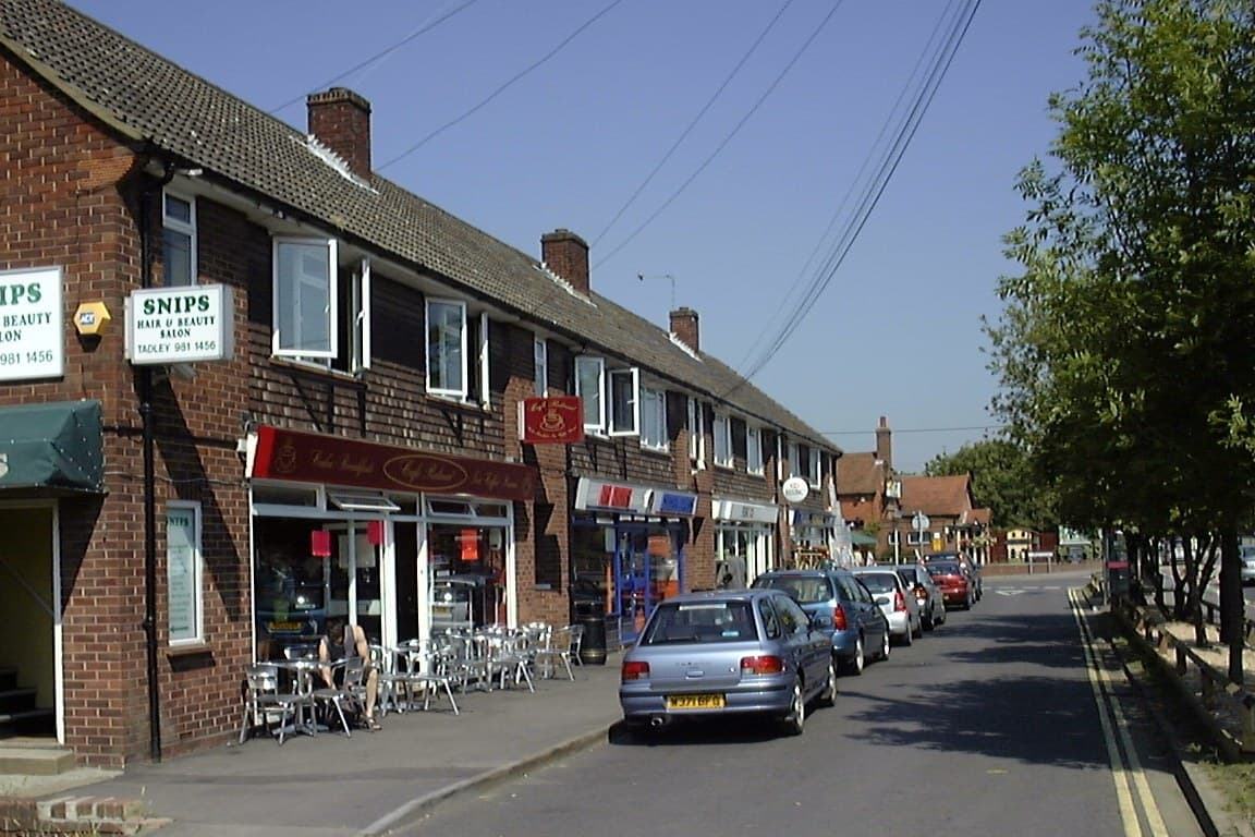 The Parade on Mulfords Hill, a row of shops with a hair salon on the corner and a cafe under a red awning, cars parked along the kerb.